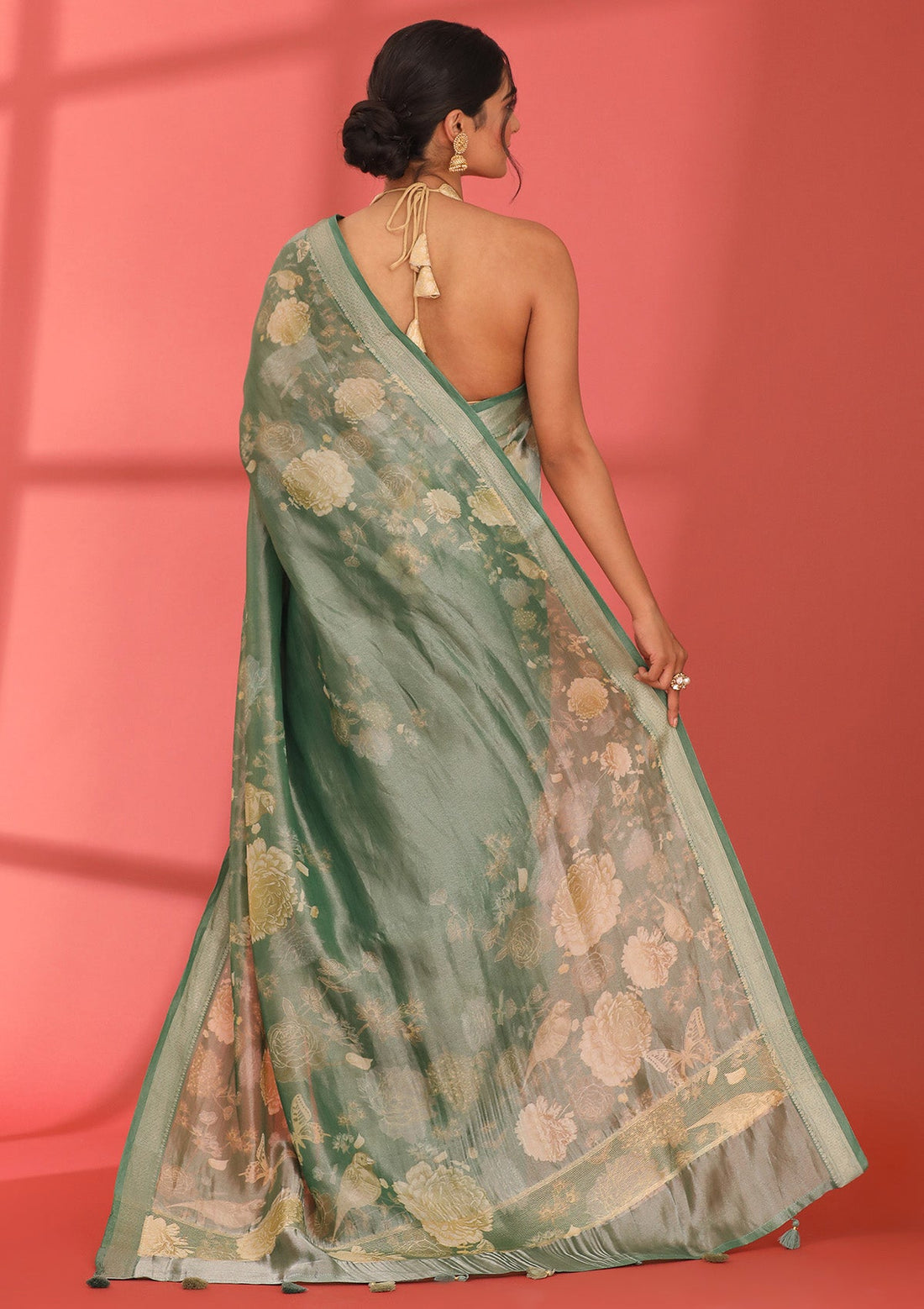 A woman shown from the back wearing a sage green saree with beige floral patterns and a matching blouse, posed against a solid coral-pink studio backdrop.